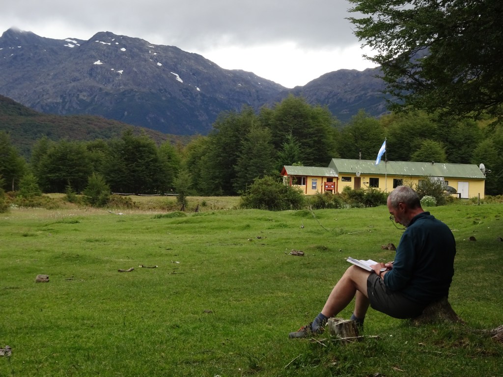 Uomo seduto su un tronco che scrive in un quaderno, circondato da un paesaggio montano e una casa gialla sullo sfondo.