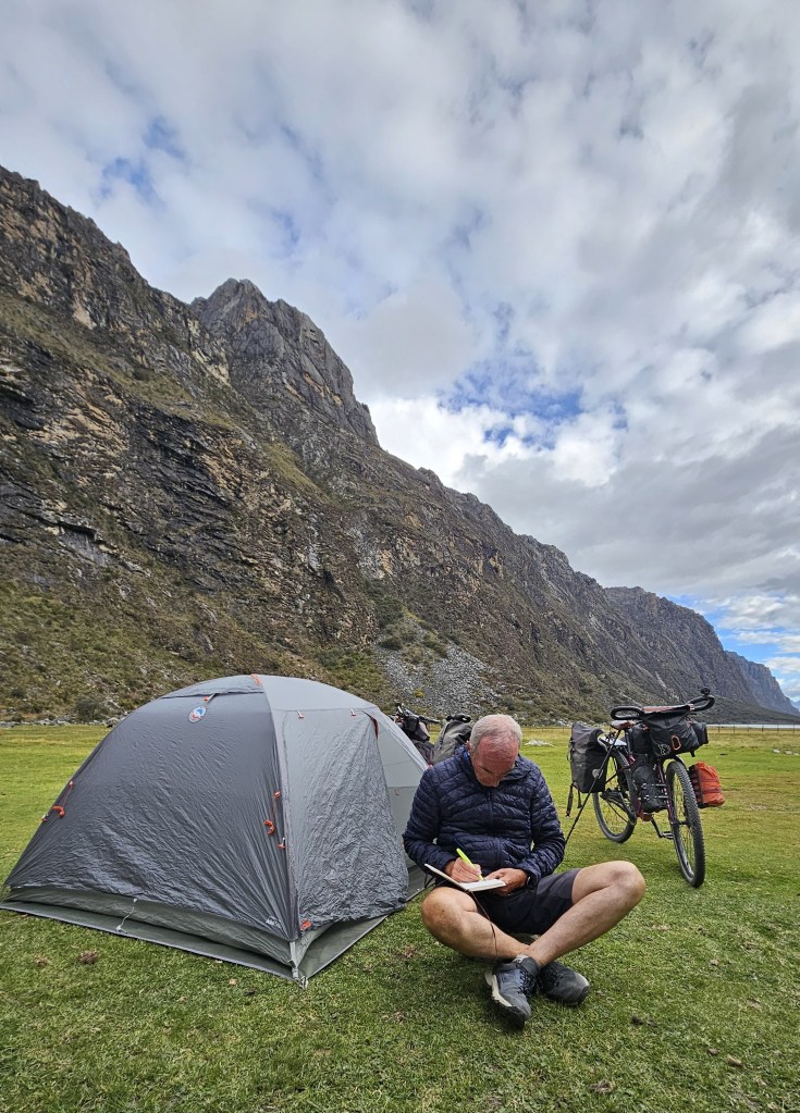 Un uomo seduto sul prato che scrive in un quaderno accanto a una tenda e una bicicletta, circondato da montagne e cielo nuvoloso.